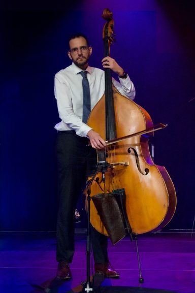 Homme en costume jouant de la contrebasse sur scène avec éclairage violet.