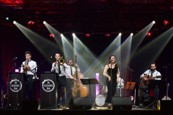 Groupe de musique sur scène, jouant avec instruments variés et chanteuse.